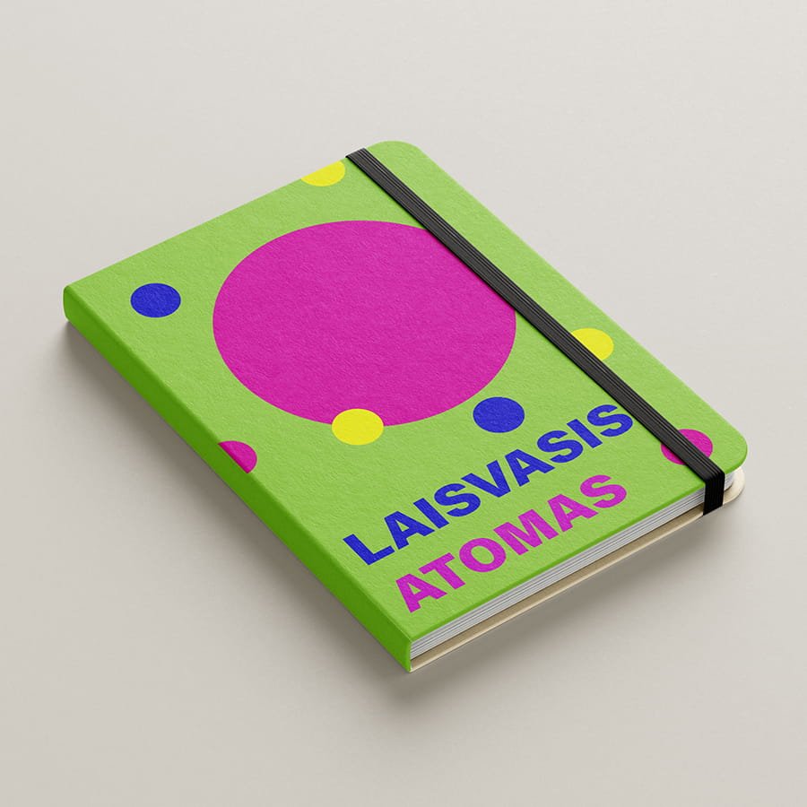 Bright green notebook cover with bold pink and blue geometric circles and the title “Laisvasis atomas,” secured with a black elastic band.