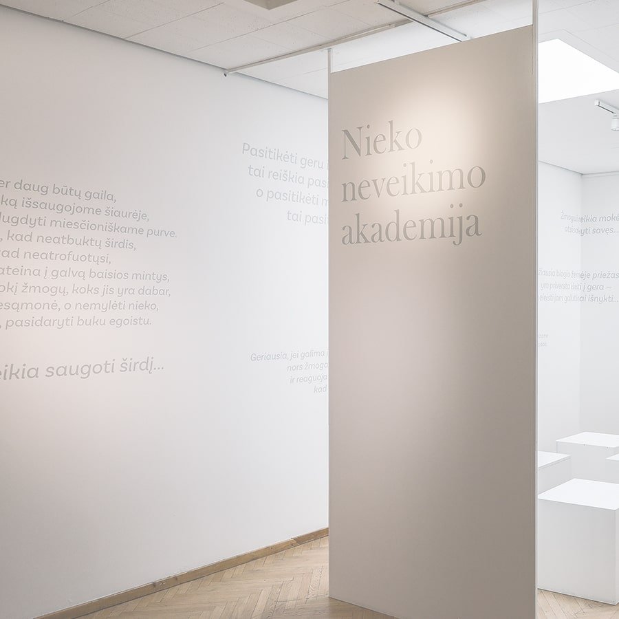 Minimalist exhibition space with soft beige panels and Lithuanian text on the walls, featuring the title “Nieko neveikimo akademija” in large elegant lettering.