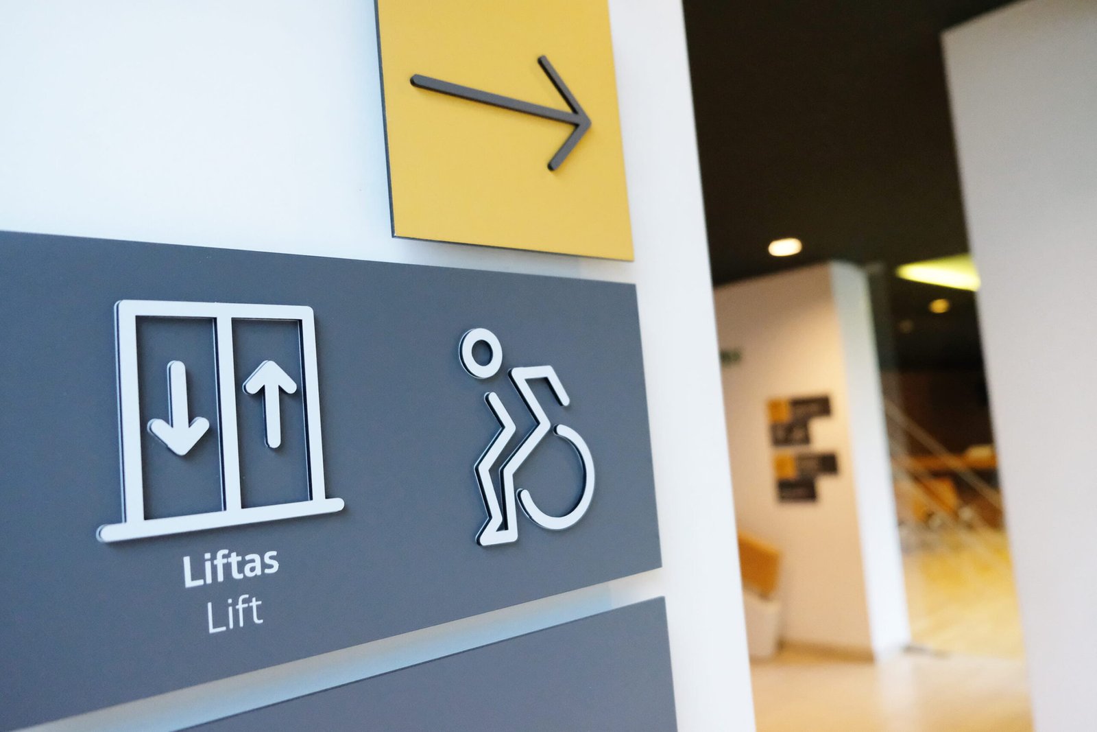 A museum wayfinding sign displays bilingual lift and wheelchair accessibility symbols with a directional arrow pointing to the right. The design uses high-contrast icons, clear typography, and a simple color palette to support accessible navigation.