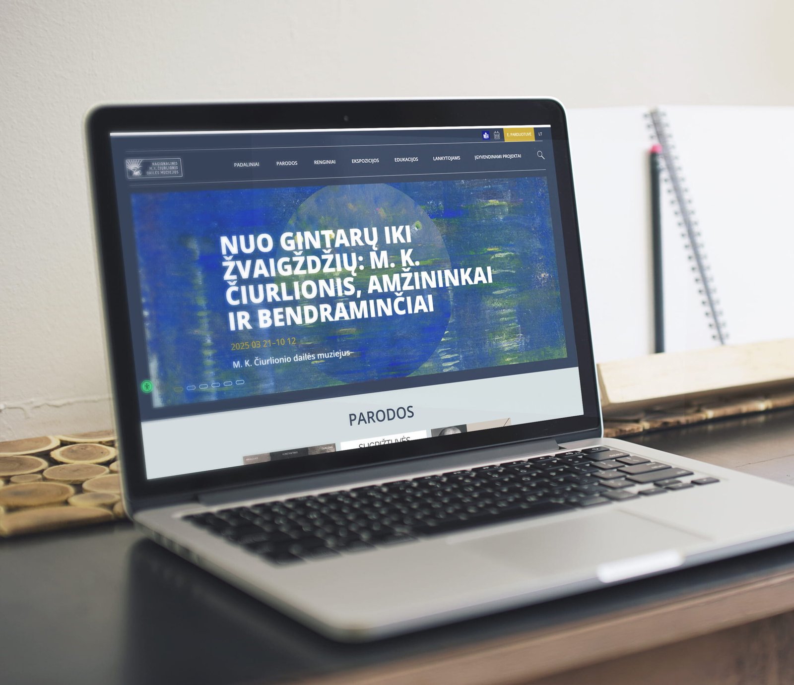 A laptop on a desk displays the Čiurlionis Museum website, featuring a large exhibition banner and the museum's homepage. The design uses bold typography, immersive artwork, and a clean, accessible layout with dark navigation and spacious content sections.