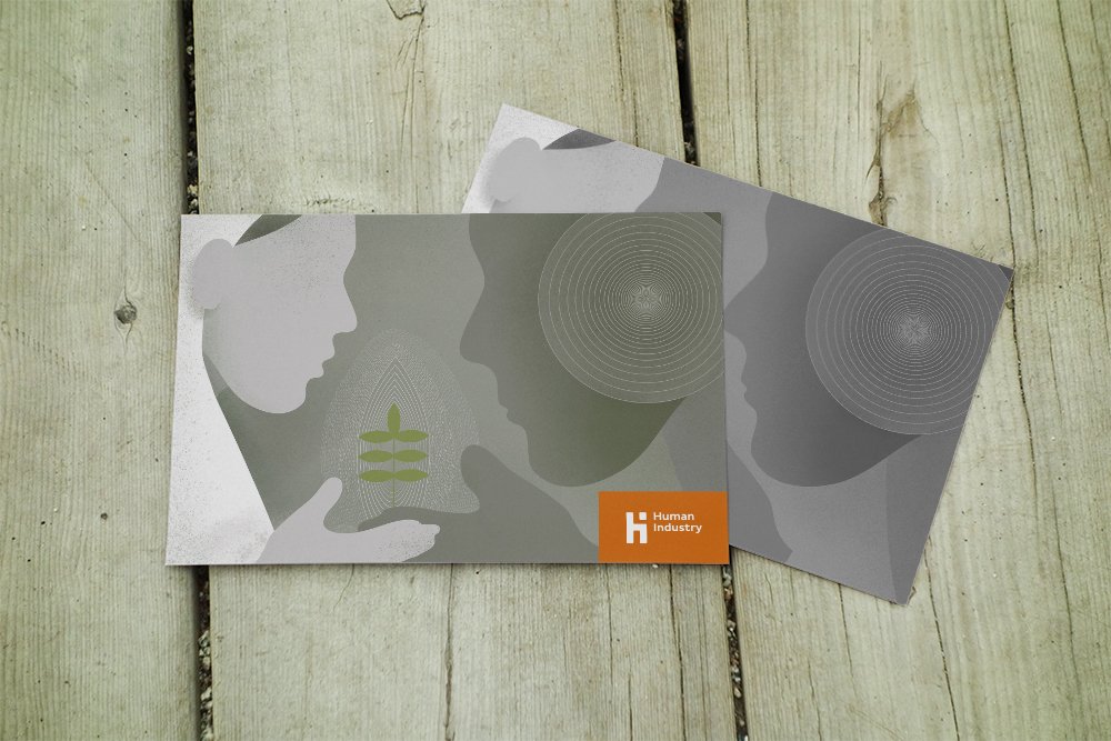 Two Human Industry printed cards lie on a pale wooden surface. The top card features two abstract profiles facing each other, with an open hand holding a small green plant between them and fine concentric patterns in the background. A bright orange block contains the white Human Industry logo. A second card with a related grey geometric illustration is partly visible underneath.