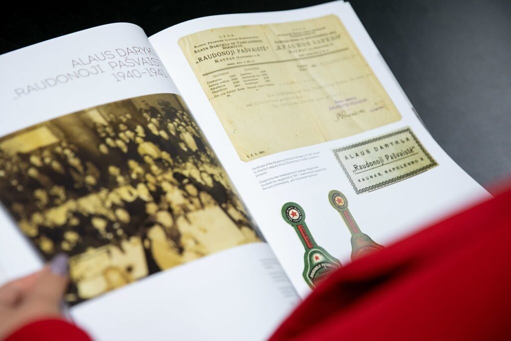 An open book displays archival photographs, documents, labels, and bottle tags from the “Red Poppy” beer hall in Kaunas.