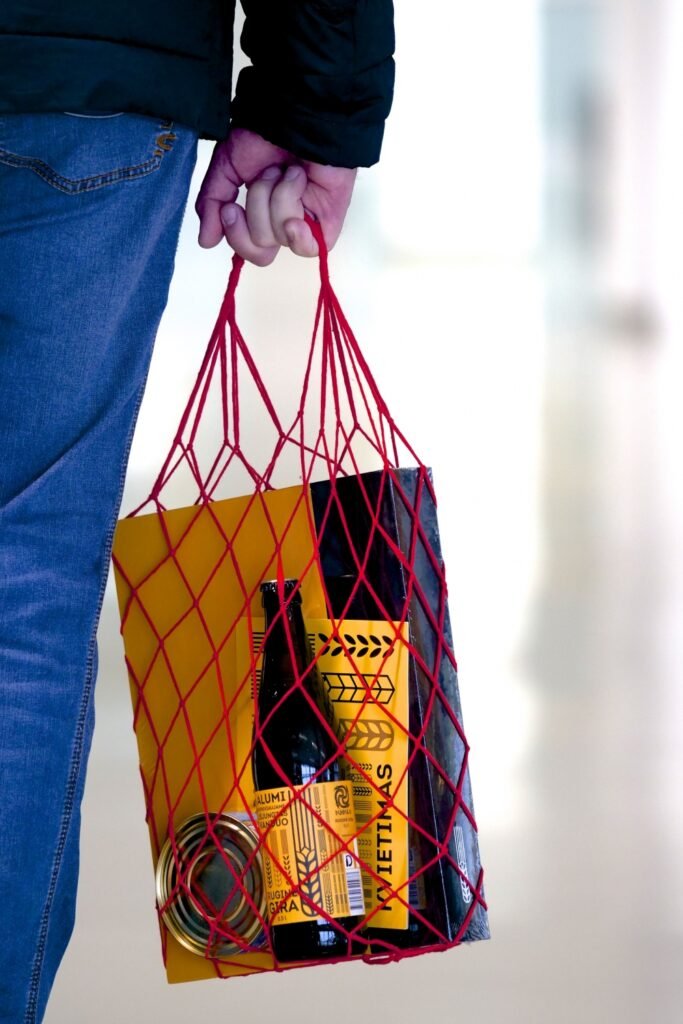 A person carries a red string bag containing a bottle, book, tin, and yellow-and-black promotional items for “No Beer for Sale — The Water’s Been Cut Off.”