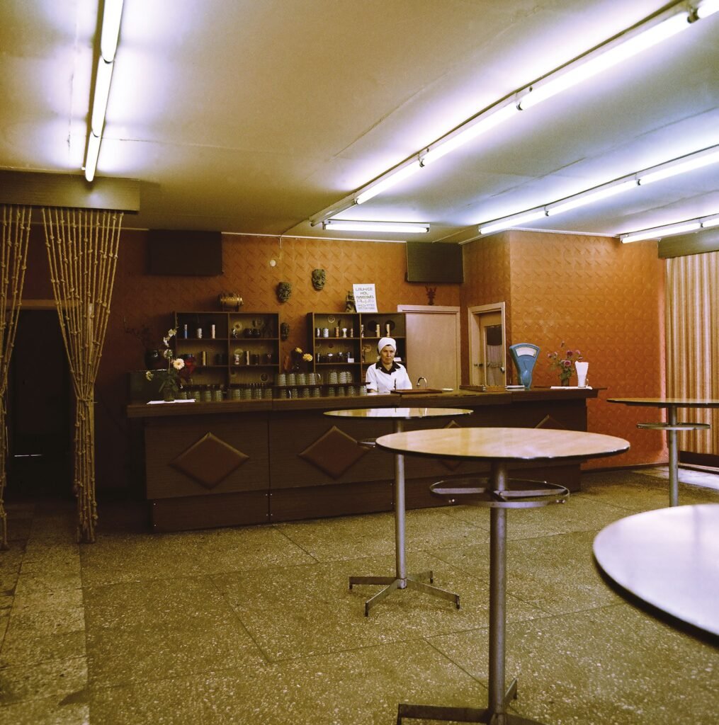 A vintage bar interior with round standing tables, patterned brown walls, fluorescent ceiling lights, and a woman behind the counter.
