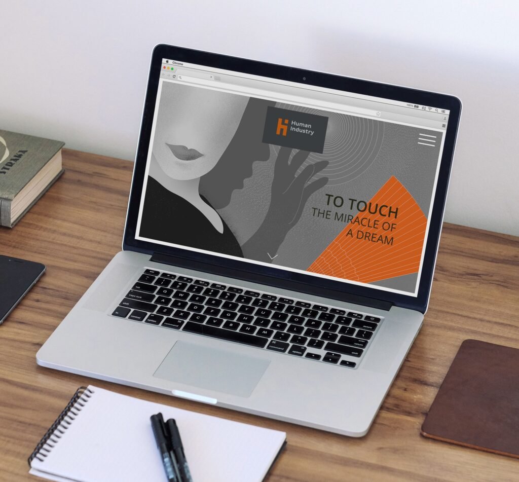 A Human Industry website is displayed on a laptop resting on a wooden desk. The homepage uses a grey and orange illustration of a stylized person reaching toward a fan of fine orange lines, with circular patterns in the background. The Human Industry logo appears at the top alongside a menu icon. Large text on the page reads: “TO TOUCH THE MIRACLE OF A DREAM.”
