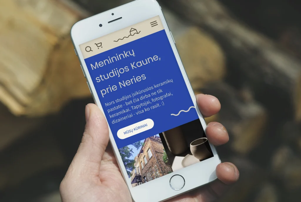 A hand holds a smartphone displaying the “Kita Neries pusė” website. A bold blue section introduces “Menininkų studijos Kaune, prie Neries” (“Artists’ studios in Kaunas, by the Neris”), explaining that the former ceramics building is now home to ceramicists, painters, photographers, designers and other artists. A “Mūsų kūriniai” (“Our works”) button appears above photographs of the brick studio building and handmade ceramics.