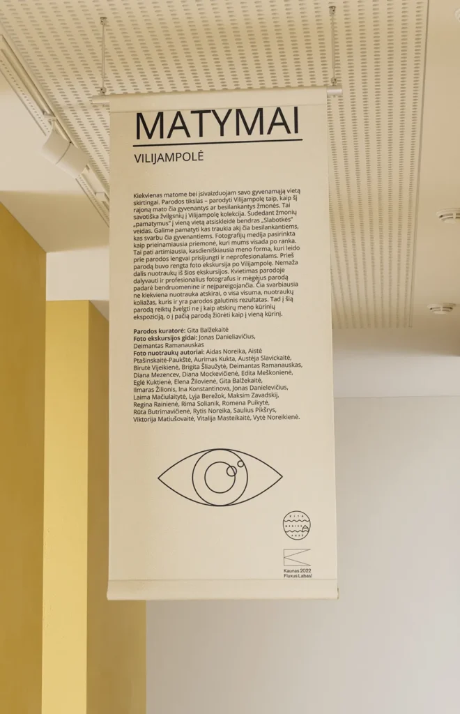 A tall cream-colored banner hangs in a bright gallery space. The heading reads “MATYMAI. VILIJAMPOLĖ,” followed by Lithuanian text explaining the community photography project, its photo excursion and exhibition, and listing the curators, excursion guide, and participating photographers. A simple line drawing of an eye and the “Kita Neries pusė” and “Kaunas 2022 Fluxus Labas!” logos appear at the bottom.