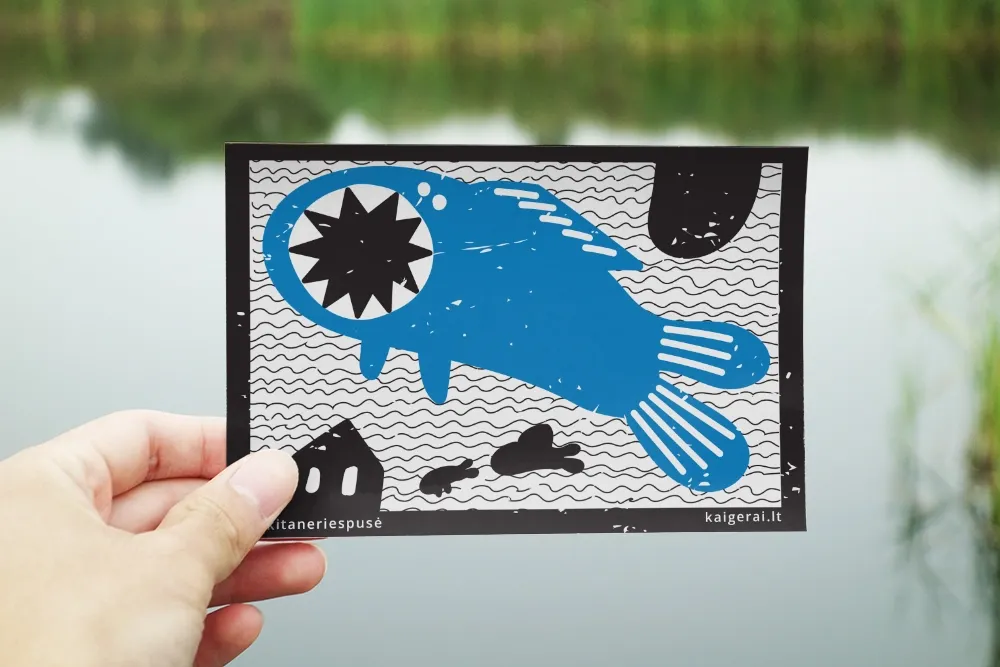 A hand holds a black, white and bright blue illustrated postcard against a blurred river background. The playful artwork shows a large imaginary blue creature surrounded by wavy lines and small black shapes. Text along the bottom reads “kitaneriespusė” and “kaigerai.lt.”