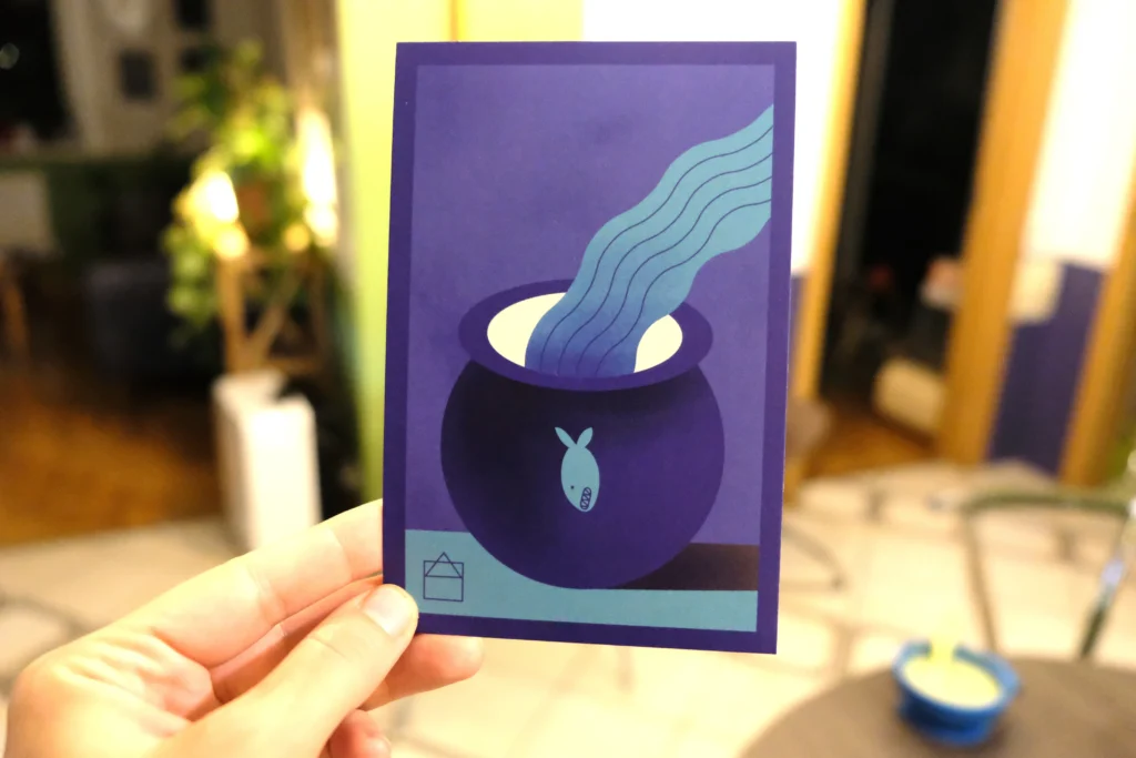 A hand holds a purple and blue postcard featuring a dark round pot with a tiny fish symbol. A stream of wavy blue water appears to pour into or rise from the pot.