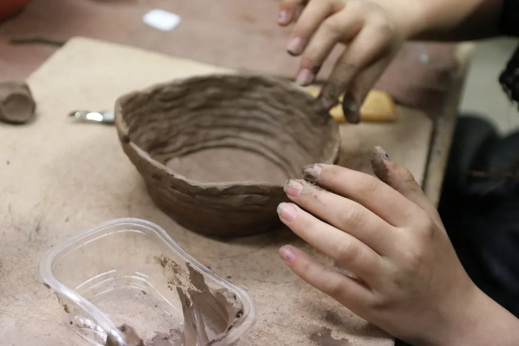 Close-up of clay-covered hands shaping a handmade ceramic bowl during a workshop. The bowl has an uneven rim and visible horizontal finger marks, giving it a wonderfully handmade character.