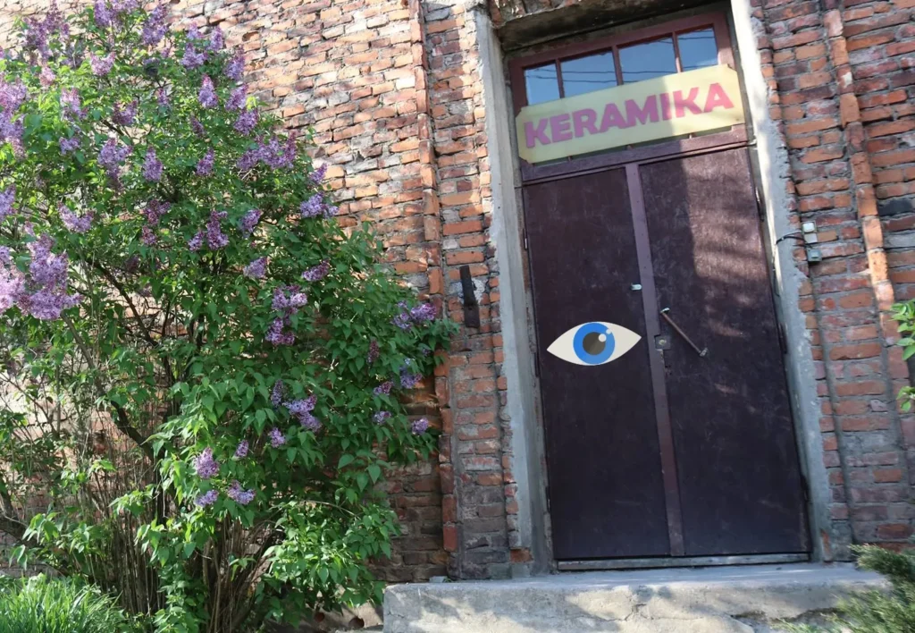 Sunlit entrance to an old red-brick building surrounded by blooming purple lilacs. Above the dark wooden doors, a sign reads “KERAMIKA.” A colorful illustrated eye symbol has been added to the door, connecting the location with the visual identity of the “Matymai” photo exhibition.