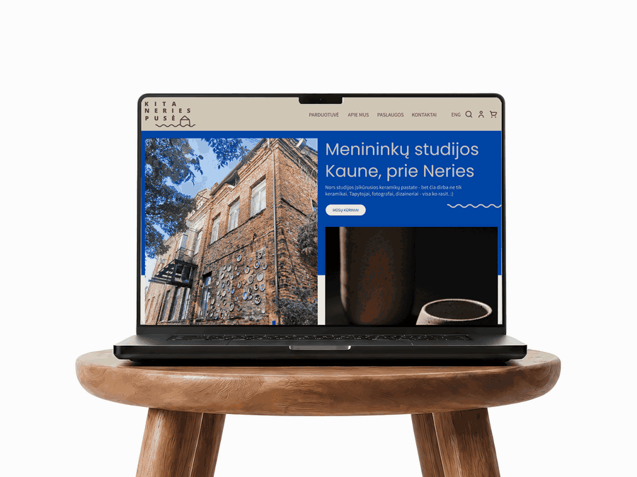 An open laptop rests on a deep-blue sofa, showing the “Kita Neries pusė” community art e-shop. The cream-and-blue homepage features the artists’ studio building, handmade ceramic vessels and the heading “Menininkų studijos Kaune, prie Neries” (“Artists’ studios in Kaunas, by the Neris”). The navigation includes Shop, About Us, Services, Contacts and English-language options.