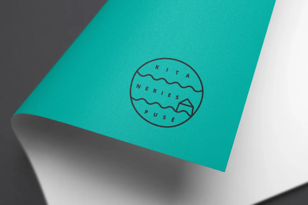 Close-up mock-up of the black “KITA NERIES PUSĖ” circular logo printed on a curved turquoise surface, featuring two wavy river lines and a small house symbol.