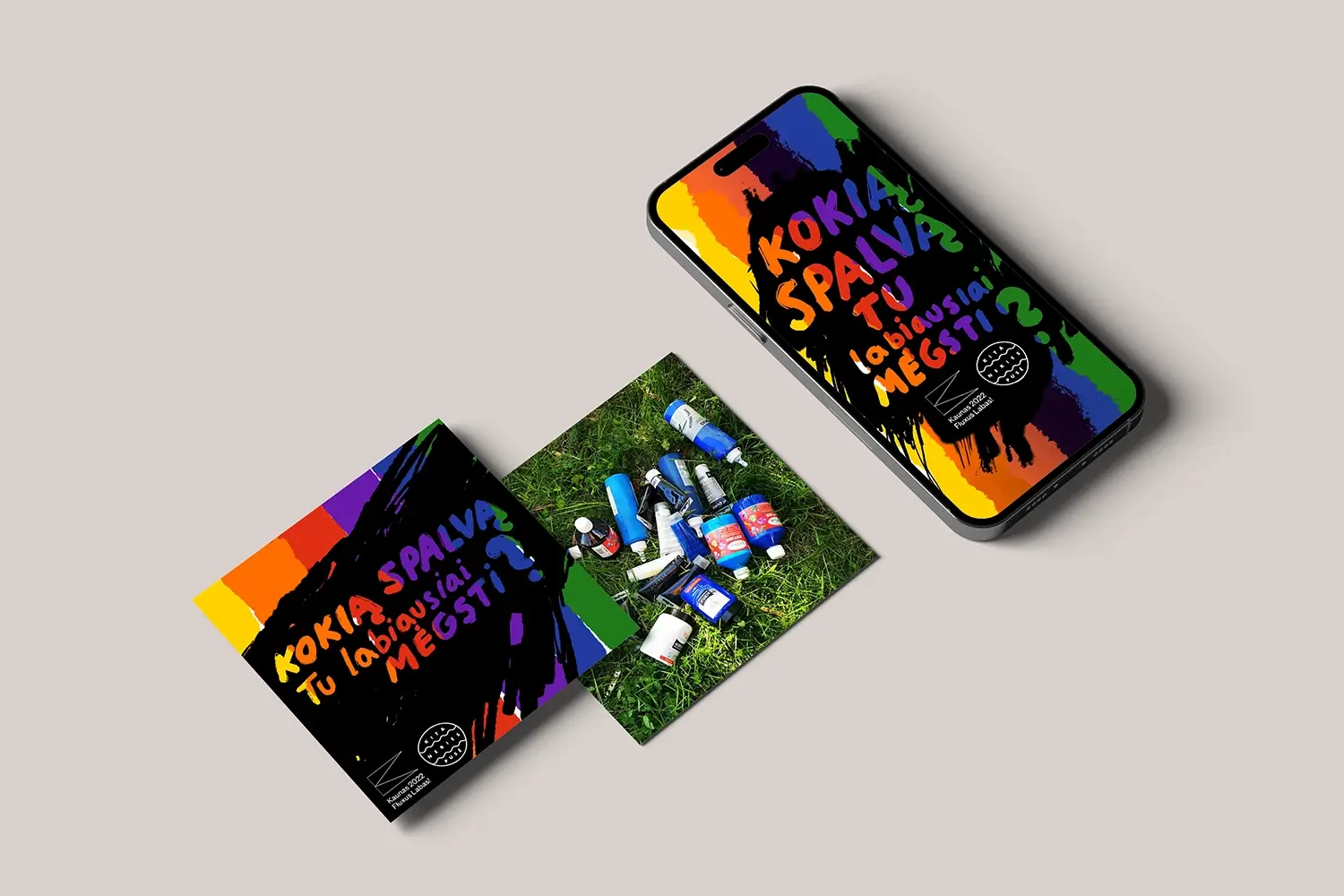 Smartphone and printed cards displaying the colorful workshop design with the Lithuanian question “KOKIĄ SPALVĄ TU LABIAUSIAI MĖGSTI?” (“What color do you like the most?”). A photograph of assorted paint bottles lying on grass is placed between the cards.