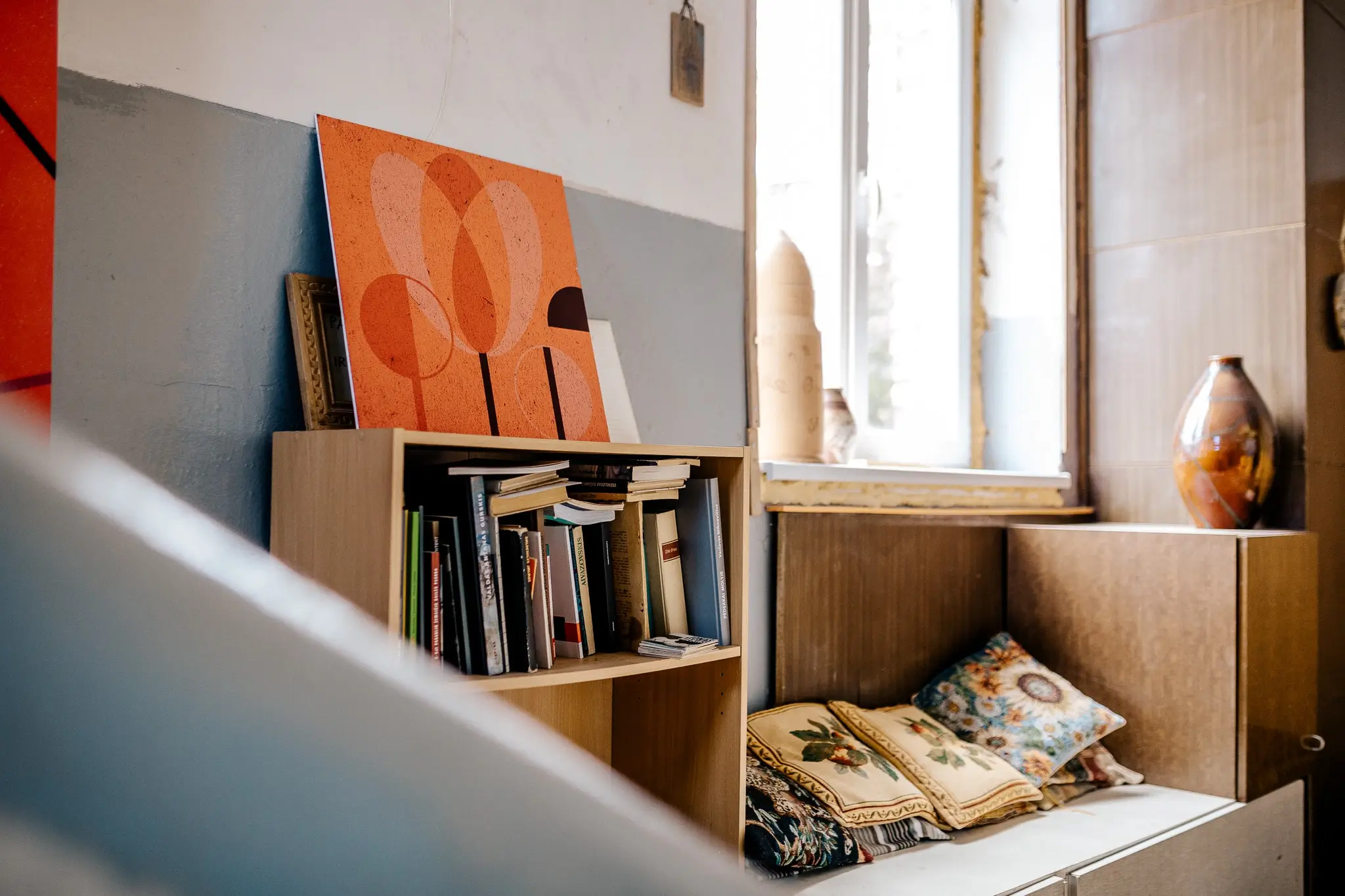 Sunlit interior with bookshelves, patterned cushions and an orange abstract artwork leaning on top of a bookcase. The artwork features overlapping translucent rounded forms in warm orange and brown tones.