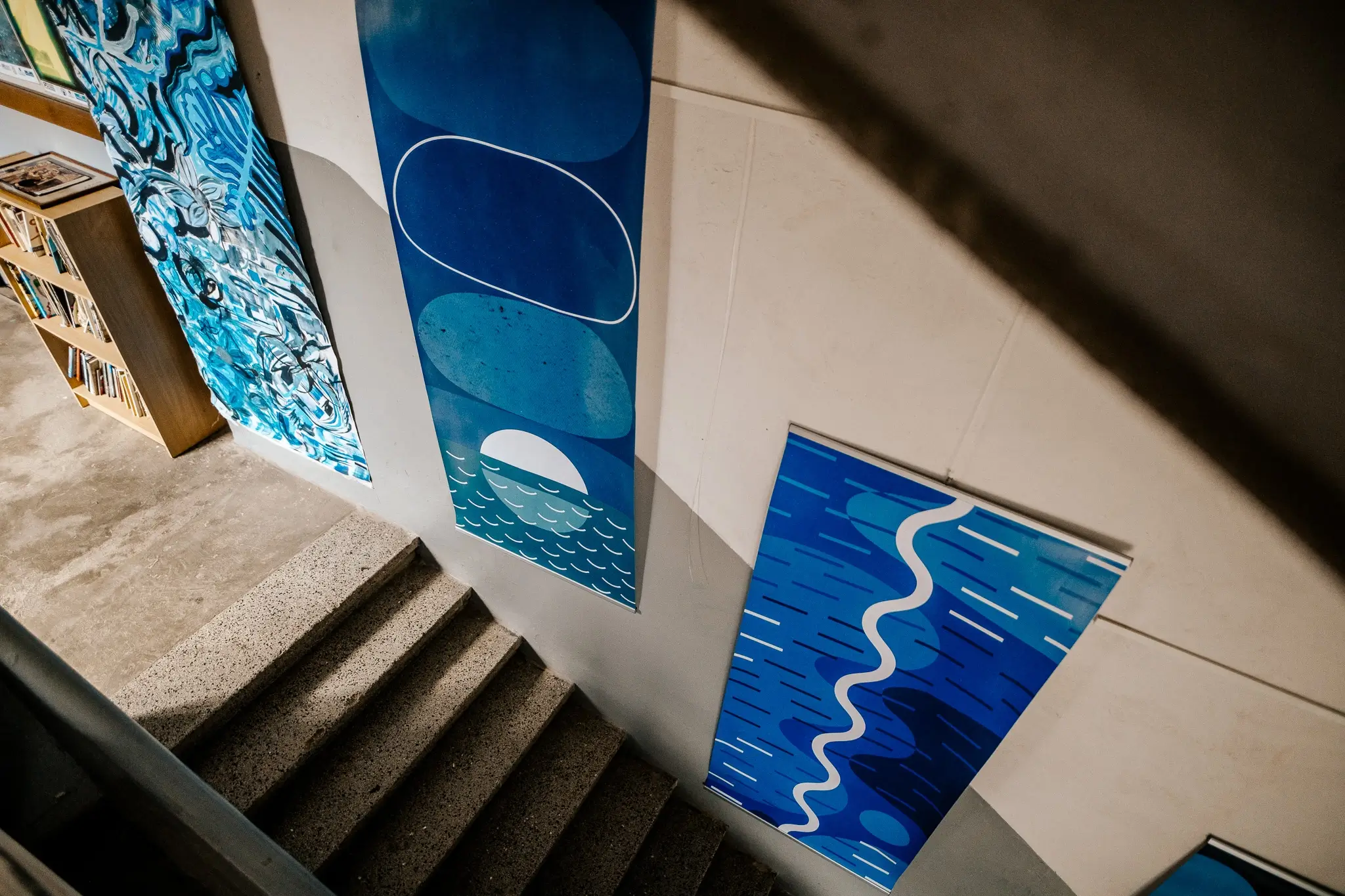 View down a concrete staircase lined with colorful artworks. Two tall blue geometric pieces feature abstract shapes, white lines, a half-circle and stylized waves.