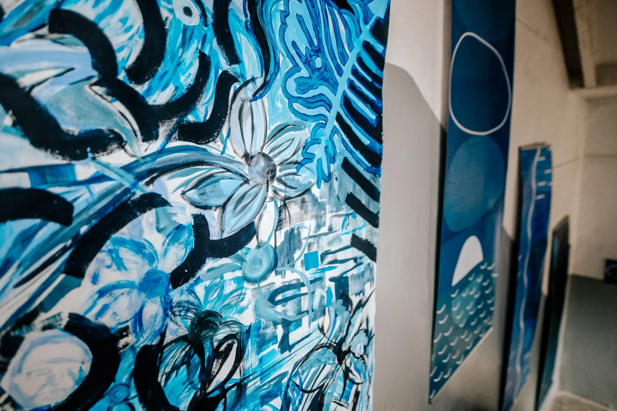 Close-up of a large expressive painting filled with blue, white and black flowers, leaves and sweeping brushstrokes. In the background, tall blue geometric artworks hang along the wall beside a staircase.