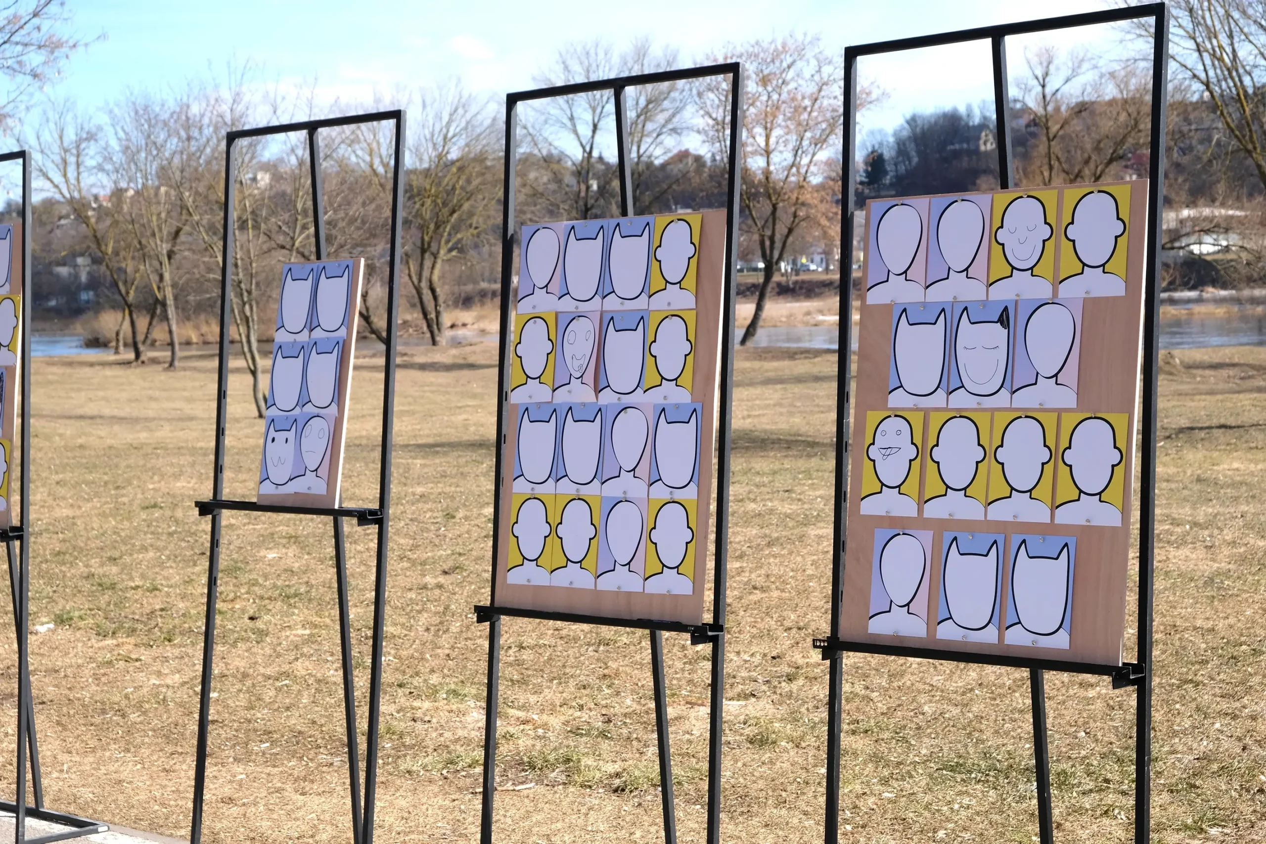 Black metal display frames stand in a park beside the river, holding grids of simple human and cat portrait templates. Some blank faces have already been transformed with hand-drawn eyes, smiles, and other playful details.