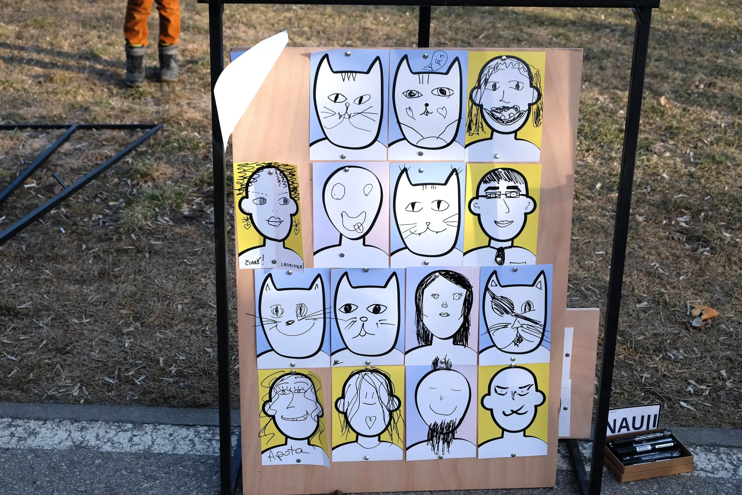 A wooden board displays sixteen hand-drawn human and cat portraits created from simple templates. Participants have filled them with expressive faces, hairstyles, glasses, whiskers, moustaches, and plenty of quirky personality.