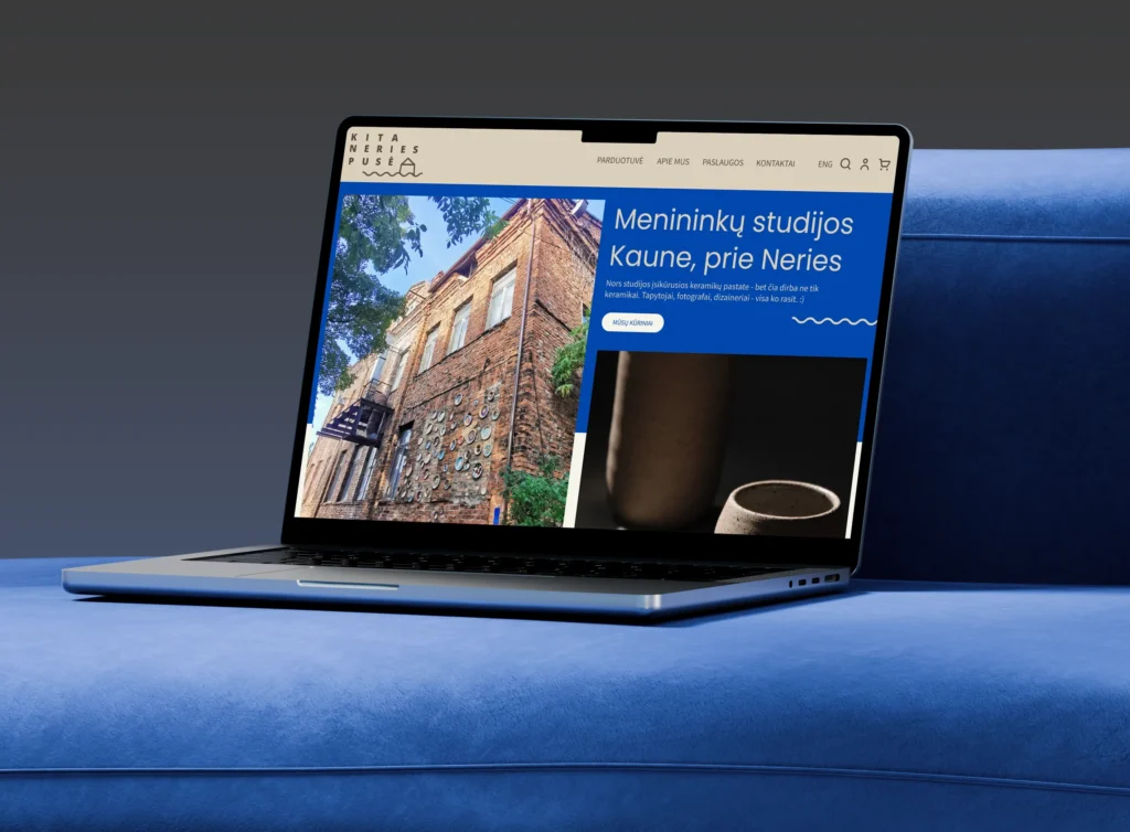 A laptop on a wooden stool displays the “Kita Neries pusė” website. The homepage combines a photograph of an old brick artists’ studio decorated with ceramic pieces and a close-up of handmade pottery. A cobalt-blue panel reads “Menininkų studijos Kaune, prie Neries” (“Artists’ studios in Kaunas, by the Neris”), introducing the local creative community.