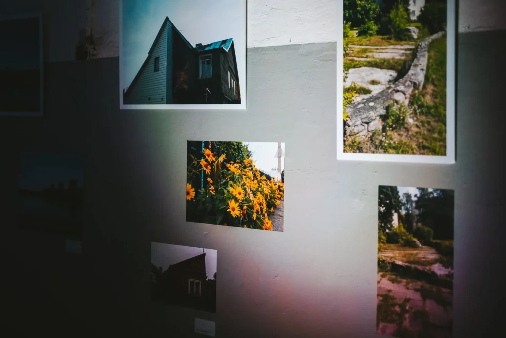 Several photographs illuminated against a pale wall show details of Vilijampolė: a sharply roofed wooden house, bright yellow flowers, an old stone boundary, another wooden building, and a sunlit path surrounded by greenery.
