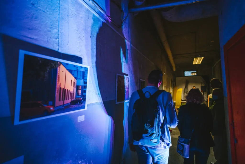 Visitors walk through a dark industrial corridor lined with community photographs. Dramatic blue and purple lighting illuminates the walls and a large architectural photograph in the foreground, giving the modest exhibition space a vivid, immersive feel.