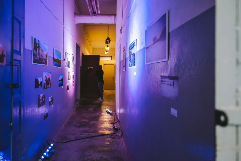 A narrow industrial corridor transformed into a photo exhibition, with photographs of Vilijampolė displayed along both walls. Blue, purple, and warm yellow lights wash over the rough walls and concrete floor, while a visitor stands near the far end of the atmospheric space.