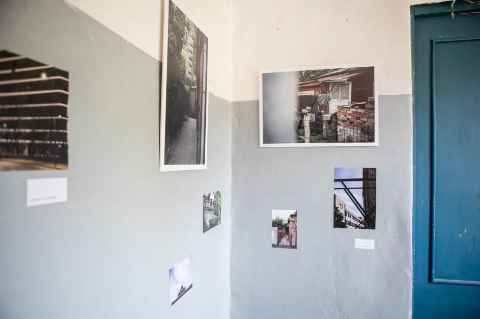 A corner of the community photo exhibition with photographs in different sizes attached directly to a pale grey wall. Images show a narrow passage, stacked bricks and old buildings, architectural details, and neighbourhood views, with a deep blue wooden door completing the modest exhibition setting.