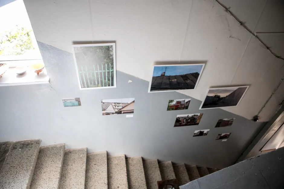 Photographs of Vilijampolė arranged informally across the white and pale grey walls of an old stairwell. The collection captures riverside views, narrow passages, wooden buildings, fences, rooftops, and other small details of the neighbourhood.