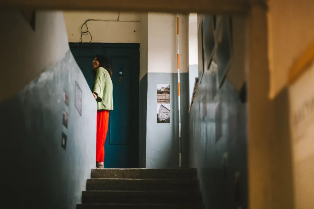 Looking up a dim staircase toward a visitor standing by a dark blue door. The visitor wears a light green top and bright red trousers, while community photographs line the stairwell walls around them.