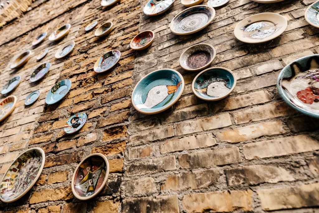 A collection of colorful handmade ceramic plates displayed across a rustic exposed-brick wall. The plates vary in size and decoration, with playful painted patterns, abstract designs, faces and animals.