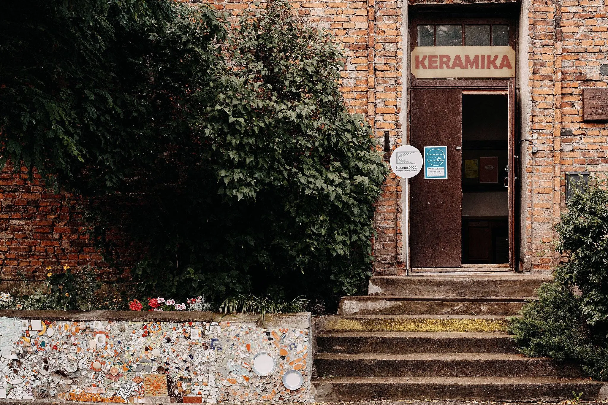 Entrance to the red-brick ceramics studio at “Kita Neries pusė,” surrounded by dense greenery and flowers. Steps lead to an open door beneath a large “KERAMIKA” sign, with a colorful ceramic mosaic decorating the low wall beside the entrance.
