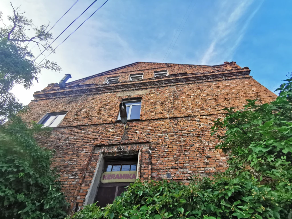 Low-angle view of an old red-brick studio building surrounded by leafy greenery under a blue sky. A sign above the entrance reads “KERAMIKA.”