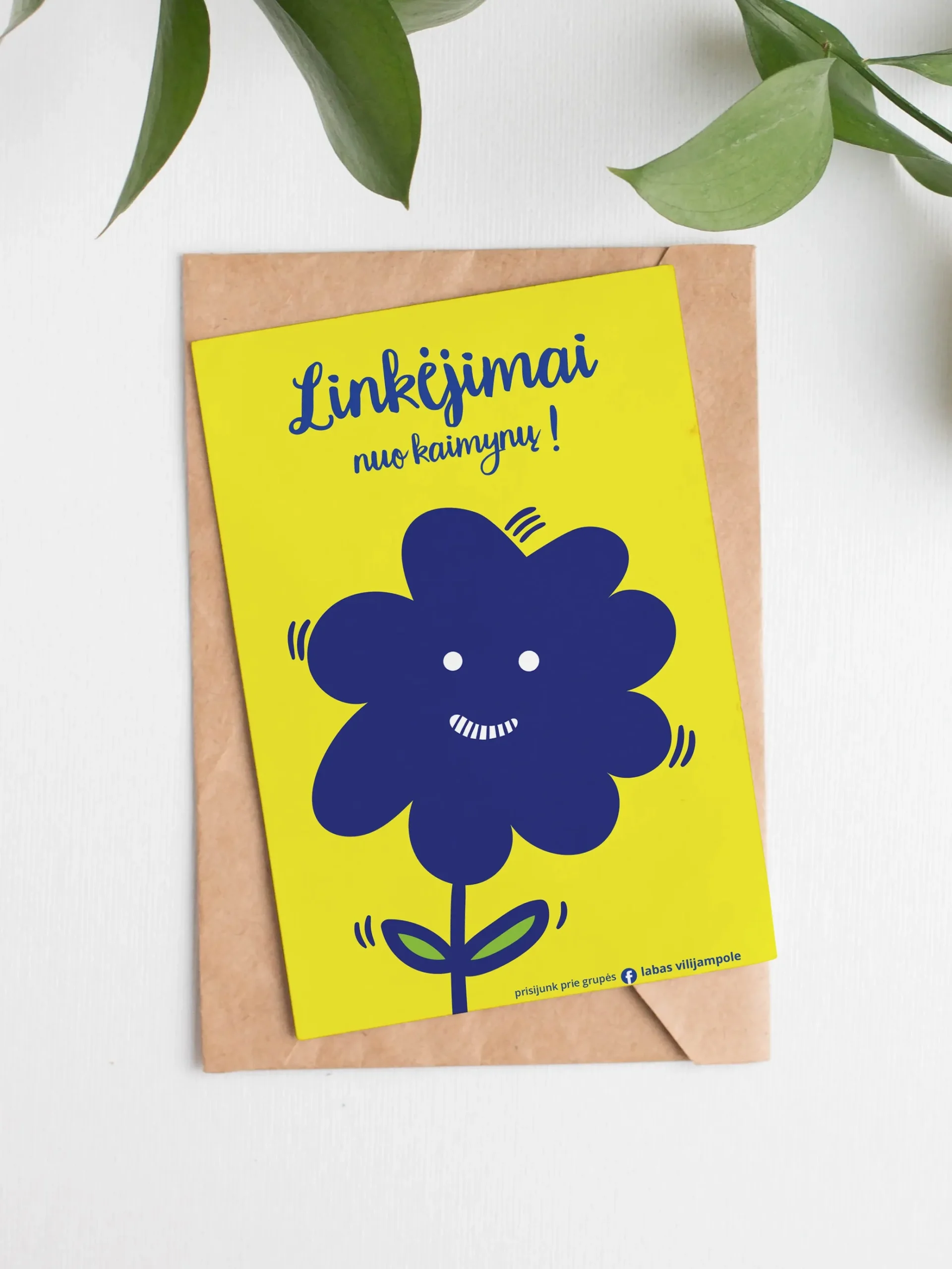 A bright yellow greeting card rests on a brown paper envelope beneath green leaves. A cheerful dark-blue flower with a smiling face fills the centre. The Lithuanian text reads “Linkėjimai nuo kaimynų!” (“Greetings from the neighbours!”), with a small invitation at the bottom to join the Facebook group “Labas Vilijampole.”