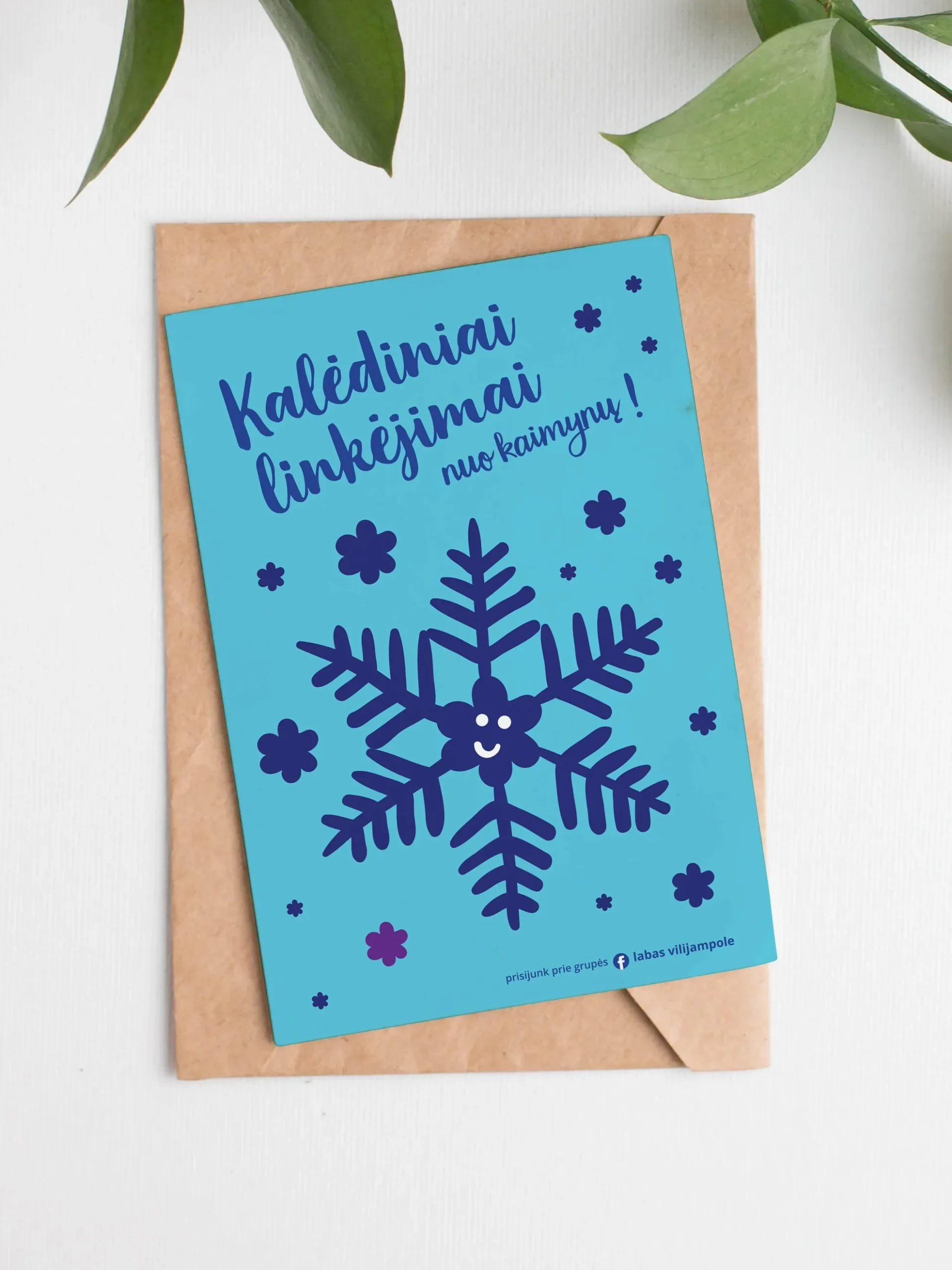 A turquoise greeting card on a brown envelope features a large dark-blue snowflake with a tiny smiling face, surrounded by smaller decorative snowflakes. The text reads “Kalėdiniai linkėjimai nuo kaimynų!” (“Christmas greetings from the neighbours!”), with an invitation to join the Facebook group “Labas Vilijampole.”