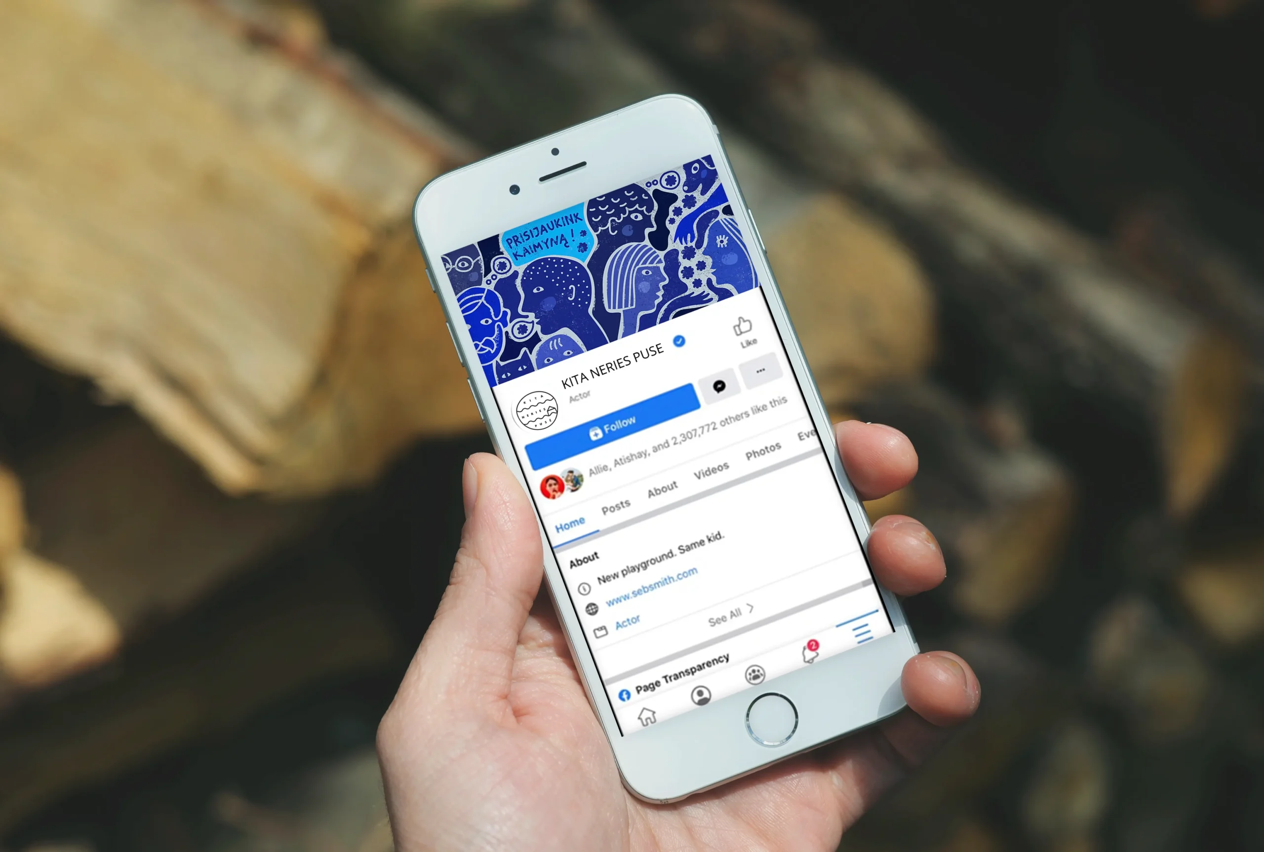 A hand holds a smartphone displaying the “Kita Neries pusė” community page. Its blue illustrated banner is filled with playful faces and neighbourhood characters and includes the message “Prisijaukink kaimyną!” (“Get to know your neighbour!”), encouraging residents to connect with one another.
