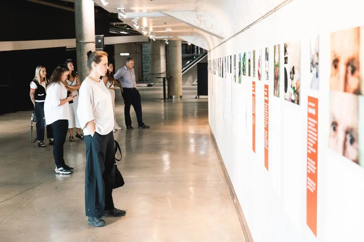 Visitors exploring a long exhibition wall featuring photographs, personal narratives and red text panels.