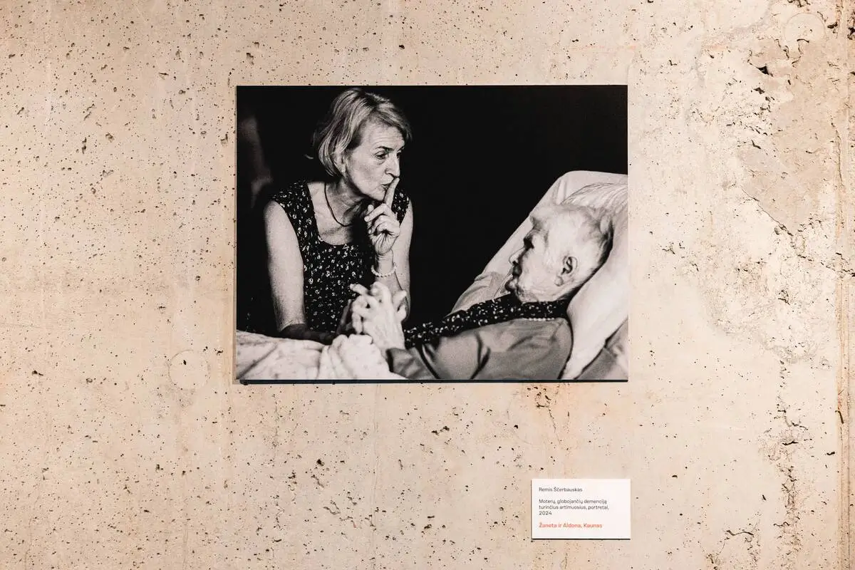 A black-and-white photograph showing a woman sitting beside an elderly person lying in bed, displayed on a concrete wall.