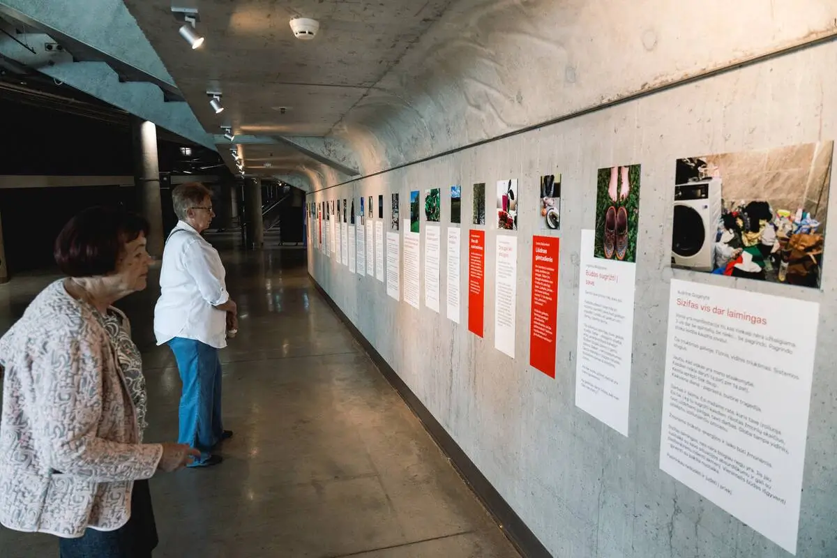 Two women viewing photographs and personal stories displayed along a concrete wall at the exhibition.