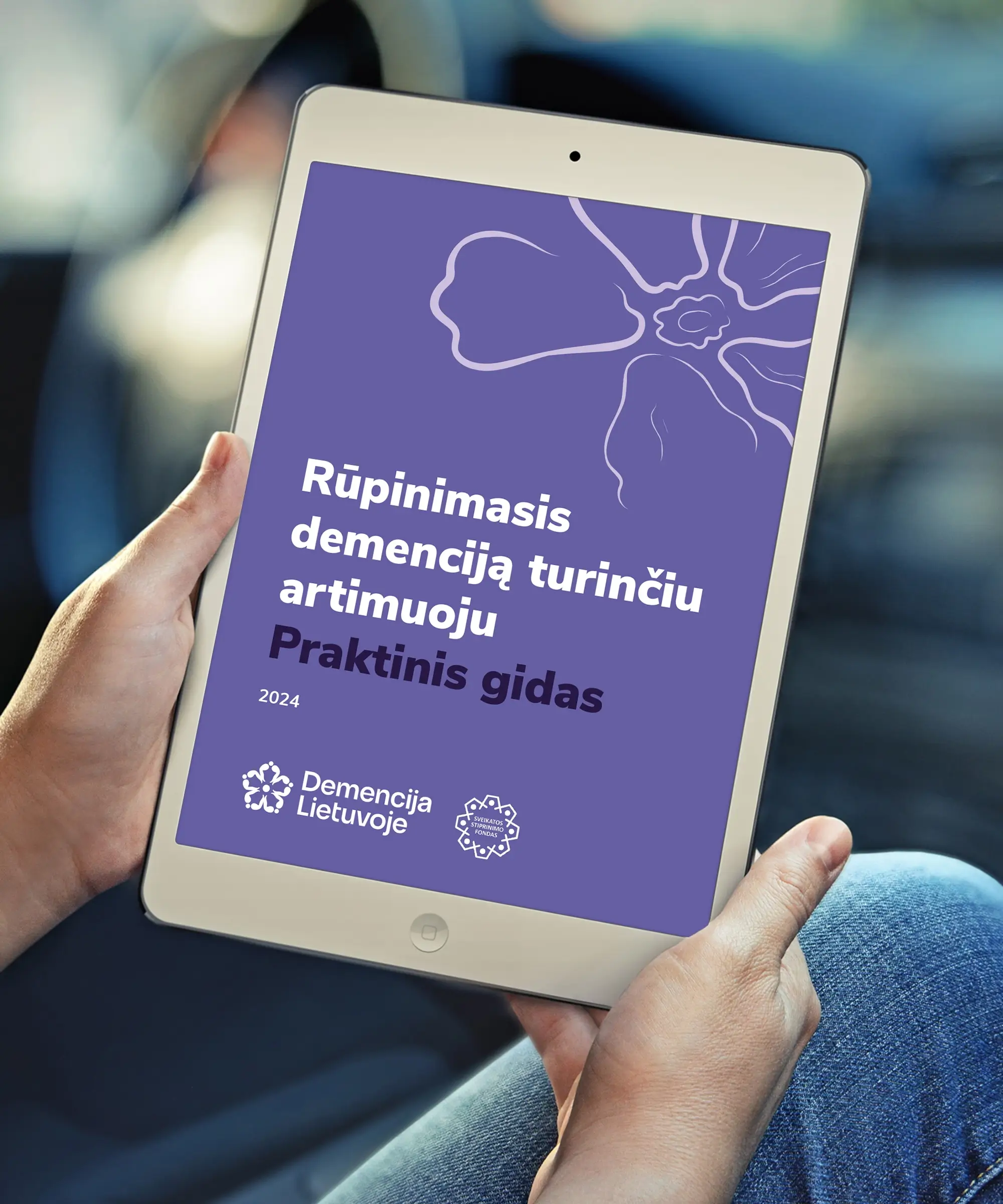A tablet displaying the purple cover of “Caring for a Loved One with Dementia: A Practical Guide,” featuring a minimalist floral line illustration and the Dementia Lithuania logo.